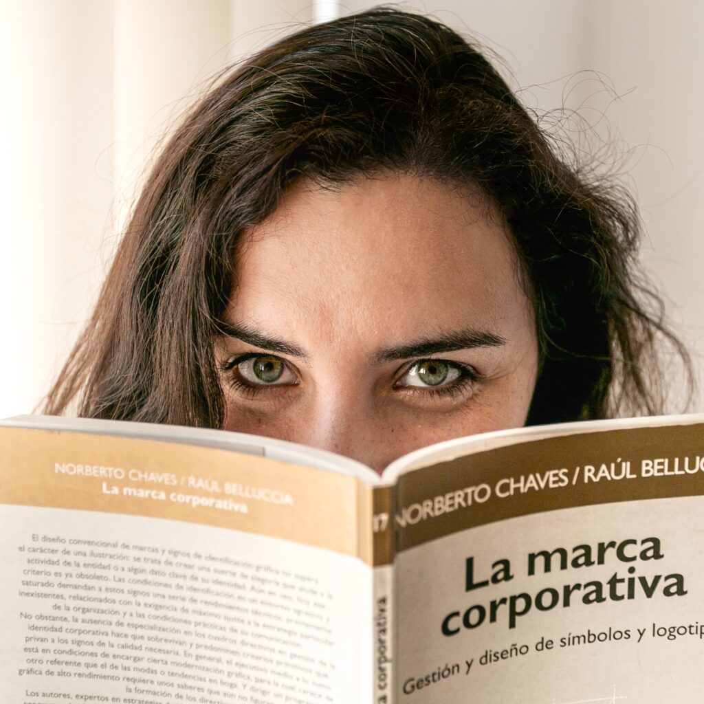 María Belén Kippes, diseñadora de marcas, argentina y fundadora de Arriver Design Studio, leyendo el libro “La marca corporativa” de Norberto Chaves y Raúl Belluccia, con la mirada asomando por encima del ejemplar, en una escena que transmite interés profesional por el branding y la identidad corporativa. María Belén Kippes, argentinian brand designer and founder of Arriver Design Studio, reading the book “La marca corporativa” by Norberto Chaves and Raúl Belluccia, with their eyes visible above the cover, conveying professional interest in branding and corporate identity.