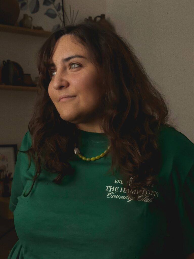 Portrait of Maria Belen Kippes, brand designer and founder at Arriver Design Studio, wearing a green “The Hamptons Country Club” shirt, a green beaded necklace, and a decorative background with pottery and leaf-patterned elements. A stylish and creative atmosphere. Retrato de Maria Belen Kippes, fundadora y diseñadora de marcas en Arriver Design Studio, con camiseta verde “The Hamptons Country Club”, collar de cuentas verdes y un fondo decorativo con cerámica y motivos de hojas. Imagen que refleja estilo personal y un ambiente creativo.